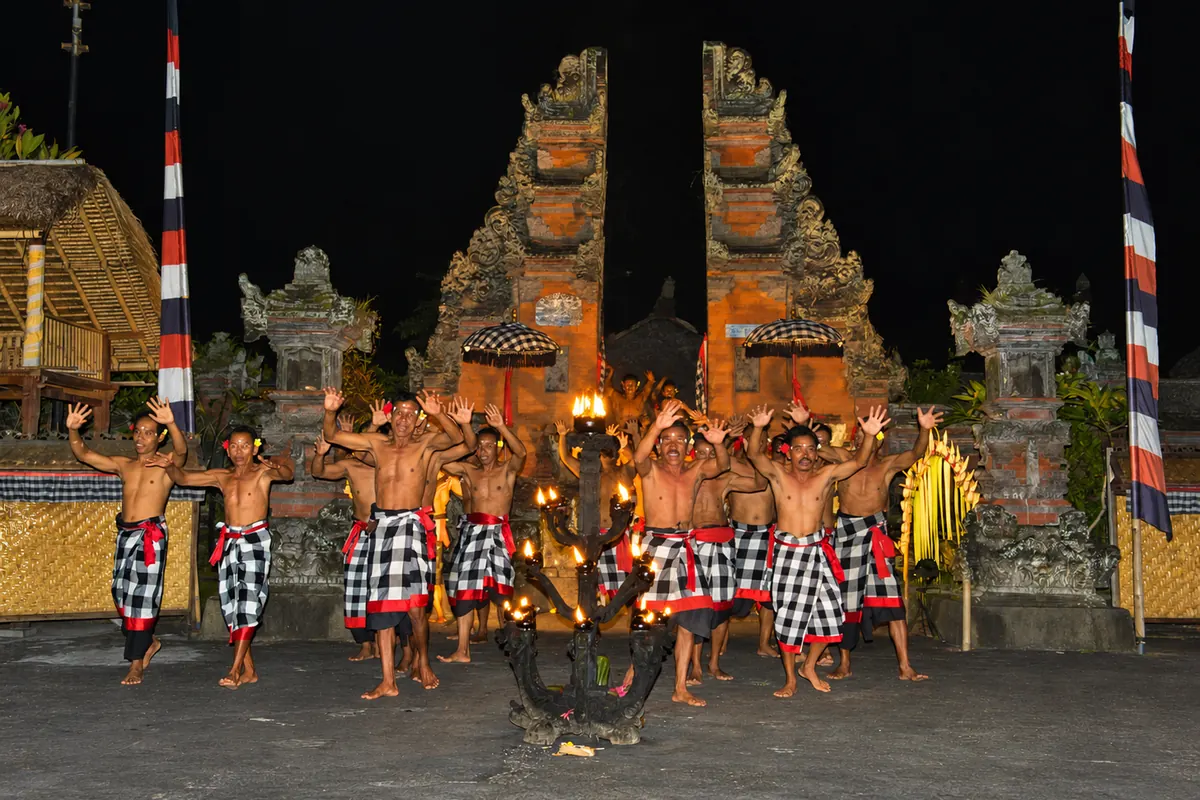 Kecak performers in checked sarongs around a flaming lamp in front of a split temple gate at Padang Tegal Kaja.
