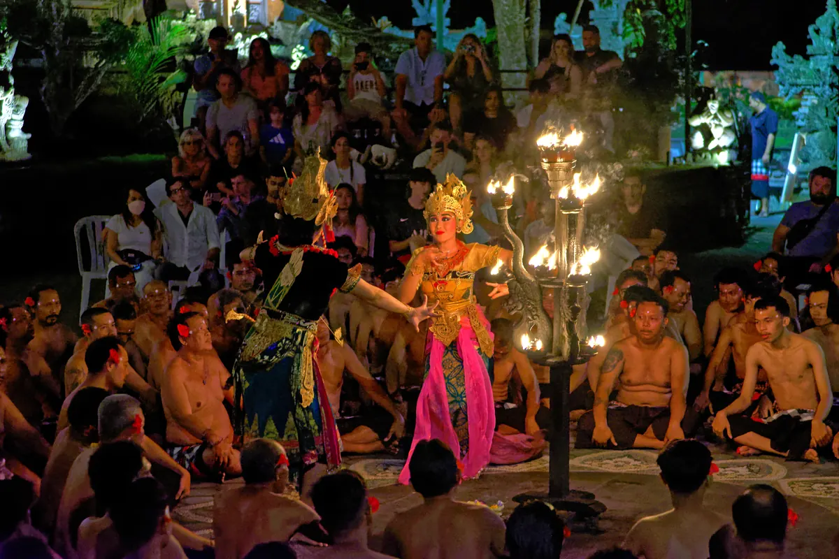 Costumed Ramayana dancers and a seated Kecak chorus beside flaming lamps at Pura Dalem Ubud's open stage.